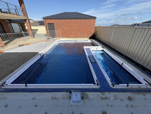 Newly installed dark blue fibreglass pool shell with attached spa/plunge pool, fitted before surrounding paving, next to a two-storey brick home