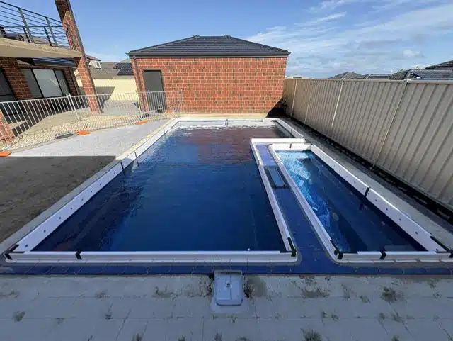 Newly installed dark blue fibreglass pool shell with attached spa/plunge pool, fitted before surrounding paving, next to a two-storey brick home